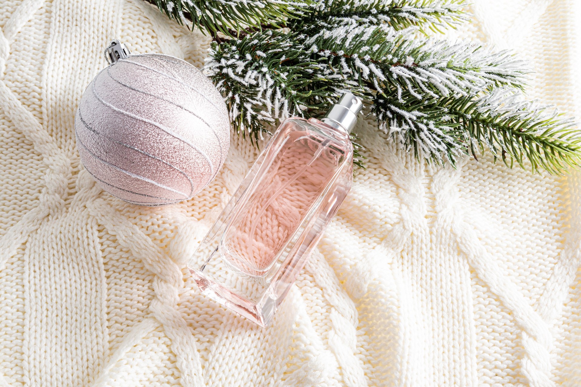 Winter New Year's advertising composition of women's perfumes on a knitted beige background with a beautiful ball and a branch of spruce.