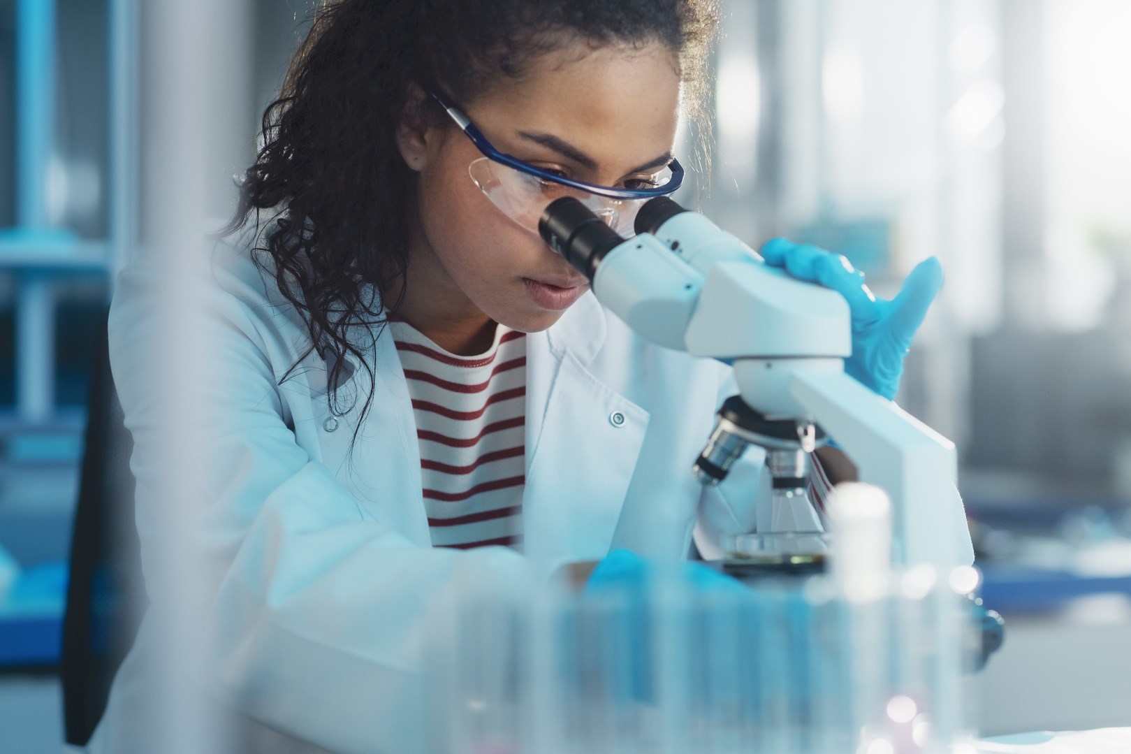 Medizinisch-wissenschaftliches Labor: Porträt einer schönen schwarzen Wissenschaftlerin, die unter dem Mikroskop eine Testprobe analysiert. Ehrgeizige junge Biotechnologie-Spezialistin, die mit moderner Ausrüstung arbeitet